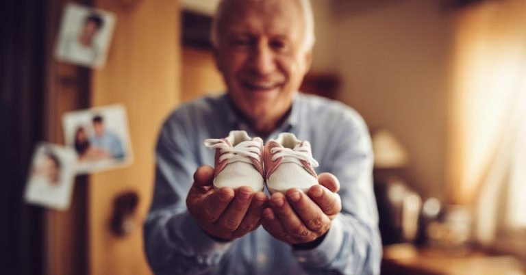 Happy grandpa-to-be holding tiny baby shoes with a warm smile, celebrating upcoming grandchild in a cozy home setting